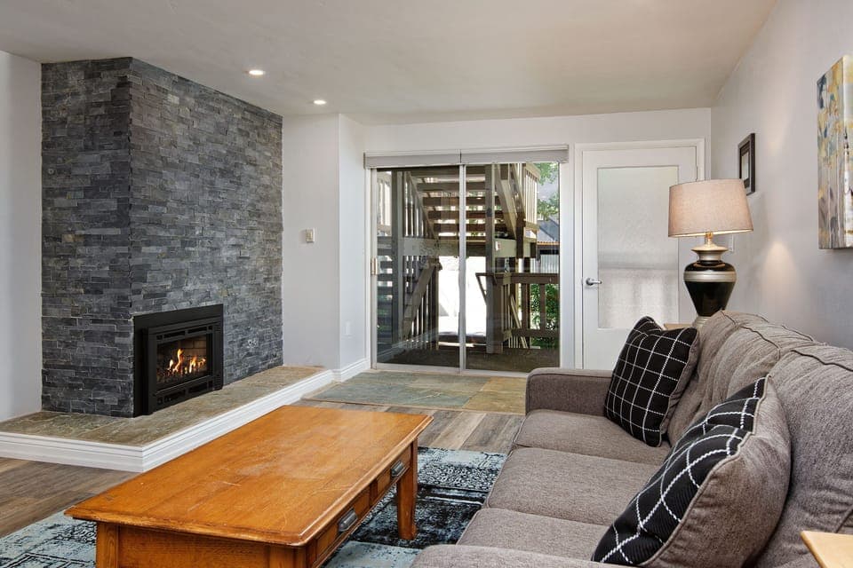 A living room with a stone fireplace, a lit fire, a gray couch with black and white pillows, a wooden coffee table, and a lamp in front of a sliding glass door leading to an outdoor area.